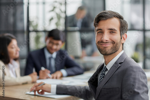 young man posing for the camera while sitting at a table in the office against the background of his colleagues