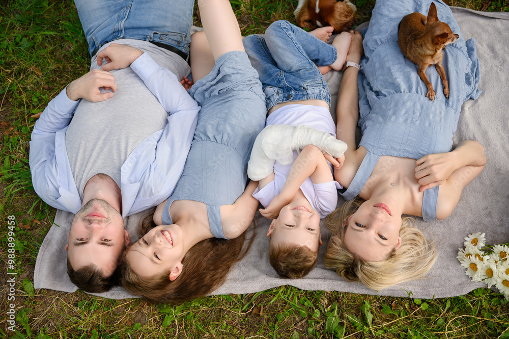 Top view of beautiful happy family lying on blanket on grass during picnic. Portrait of parents with son with plaster on arm, daughter and dogs. Family weekend concept Top view of beautiful happy family lying on blanket on grass during picnic. Portrait of parents with son with plaster on arm, daughter and dogs. Family weekend concept