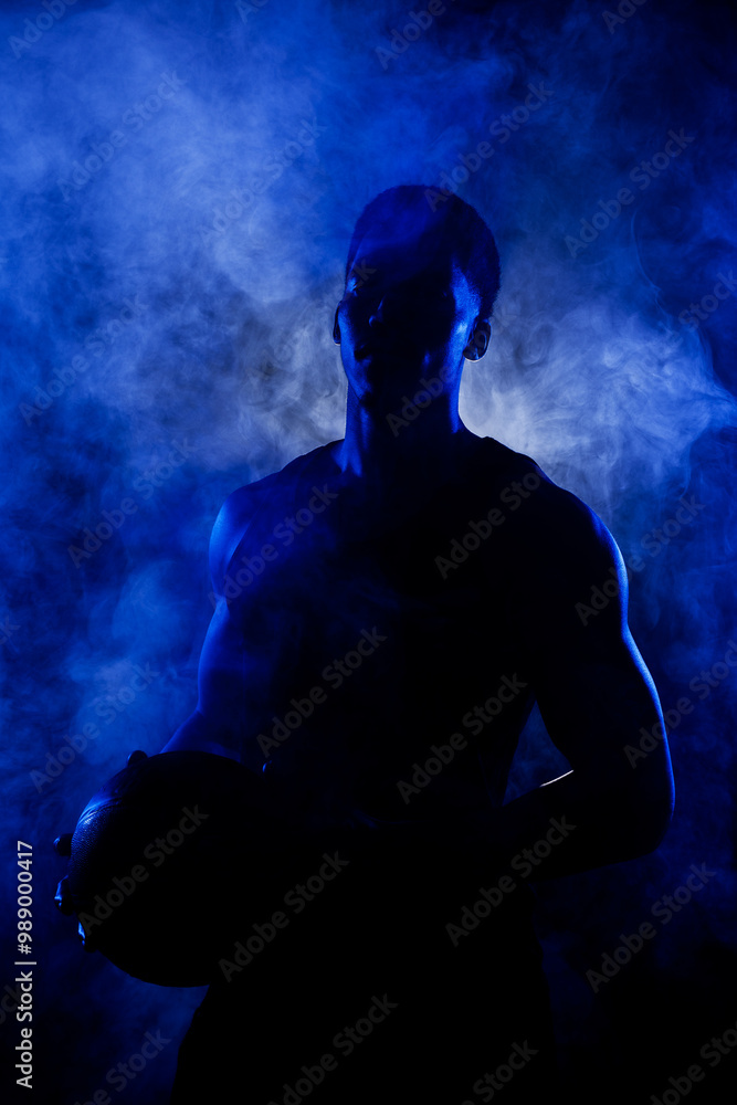 Basketball player holding a ball against blue fog background. Muscular african american man silhouette. Basketball player holding a ball against blue fog background. Muscular african american man silhouette.