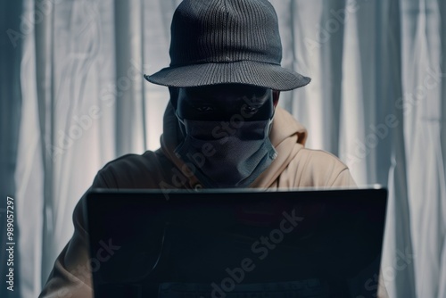 An enigmatic figure in a hat and mask sits in front of a laptop, shrouded in mystery and working in a dimly lit room. photo