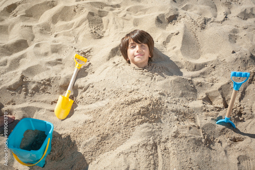 Fotografi Top view of child buried in beach sand with only head uncovered and smiling face