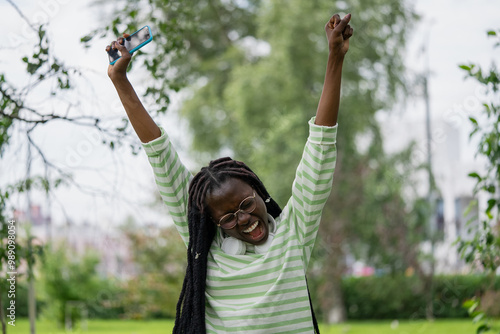 Quadro su tela African American woman joyfully raises her arms in celebration while holding smartphone, expressing excitement in green park setting