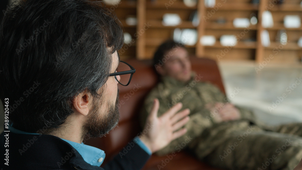 Middle aged Caucasian psychologist doctor man male speak talking to officer camouflage uniform military army soldier blurred patient guy lying on couch listening psychiatrist consultation session room Middle aged Caucasian psychologist doctor man male speak talking to officer camouflage uniform military army soldier blurred patient guy lying on couch listening psychiatrist consultation session room