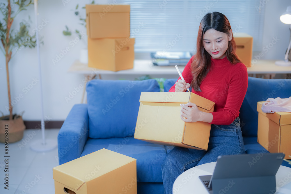 Asian woman entrepreneur prepares a package at her home office, checking orders on her tablet and writing the address on a box, focusing on excellent customer service for her online store Asian woman entrepreneur prepares a package at her home office, checking orders on her tablet and writing the address on a box, focusing on excellent customer service for her online store