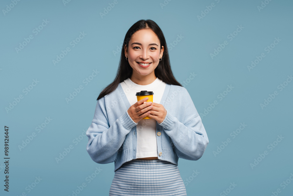 Beautiful woman holding yellow take away coffee cup, looking at camera, standing on blue background