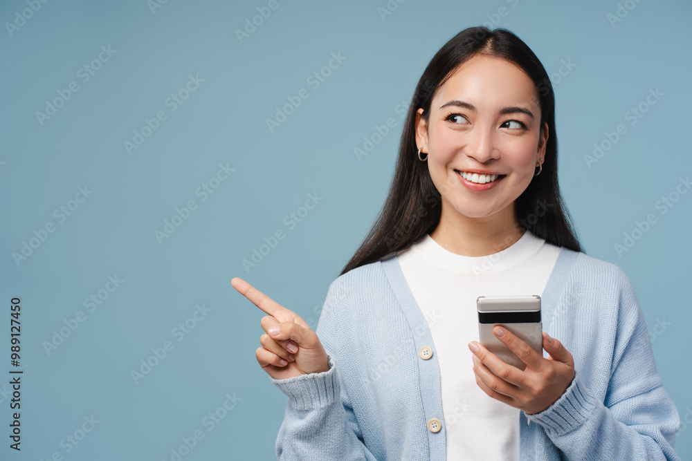 Beautiful, Asian young woman holding smartphone pointing on copy space on blue background Beautiful, Asian young woman holding smartphone pointing on copy space on blue background