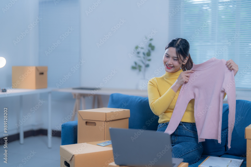 Young woman entrepreneur is broadcasting herself holding up clothing in front of a laptop, surrounded by boxes, she is smiling Young woman entrepreneur is broadcasting herself holding up clothing in front of a laptop, surrounded by boxes, she is smiling