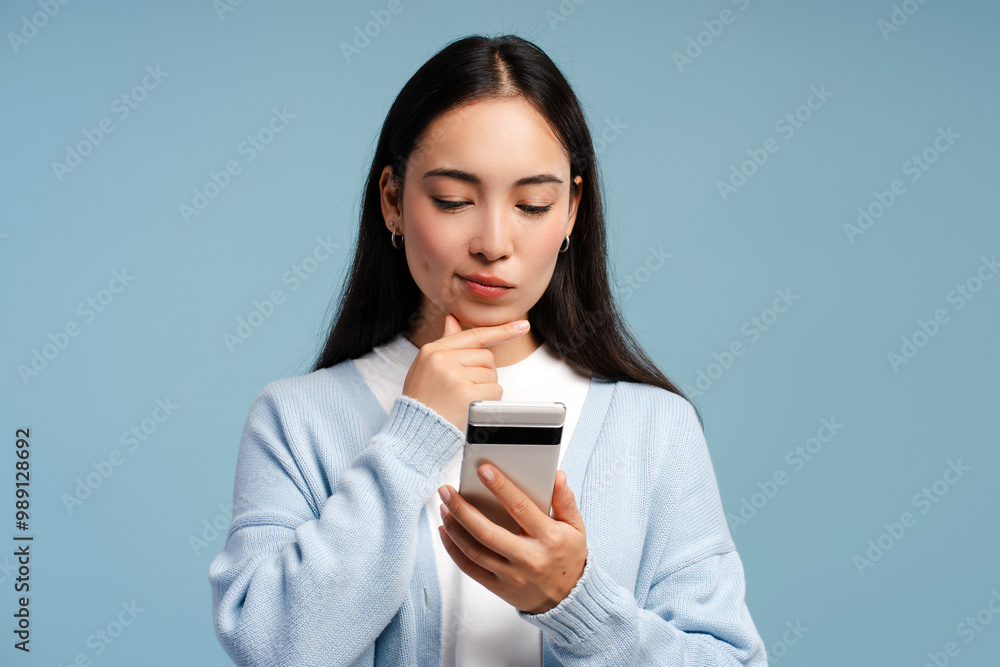 Smiling Asian woman thinking looking at smartphone, using mobile app isolated on blue background Smiling Asian woman thinking looking at smartphone, using mobile app isolated on blue background