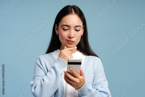 Smiling Asian woman thinking looking at smartphone, using mobile app isolated on blue background