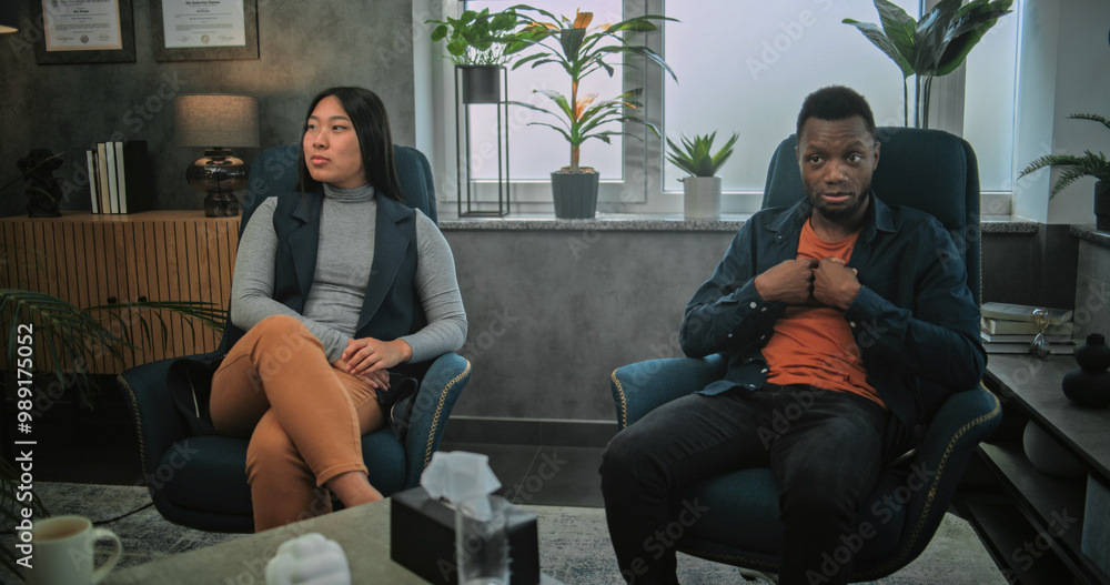 Obraz premium Back view of therapist conducting counseling session to multiethnic young family. Aggressive African American man loses temper, yells at his Asian girlfriend and starts fight during couples therapy.
