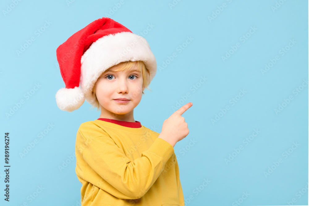 Cute boy wearing santa hat, smiling and pointing to one side on pastel blue background. Preschooler showing copy space for promotional ad. Studio portrait on pastel blue background. Cute boy wearing santa hat, smiling and pointing to one side on pastel blue background. Preschooler showing copy space for promotional ad. Studio portrait on pastel blue background.