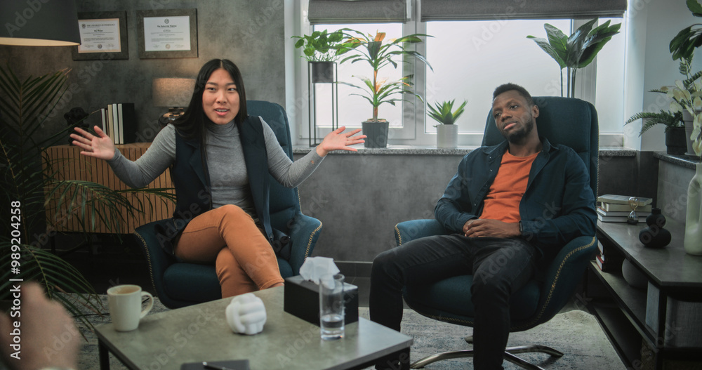 Obraz premium Back view of therapist conducting counseling session to multiethnic young family. Aggressive African American man loses temper, yells at his Asian girlfriend and starts fight during couples therapy.