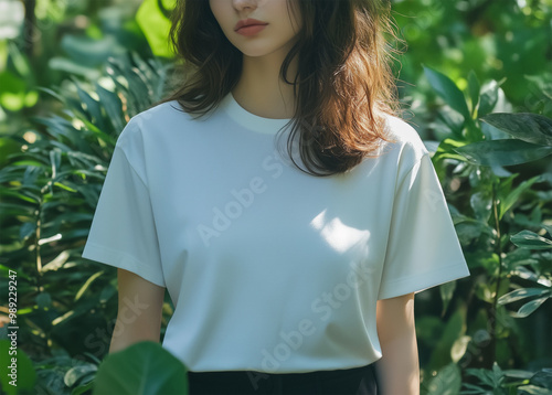 A woman in a white t-shirt stands frontally against a background of plants. photo