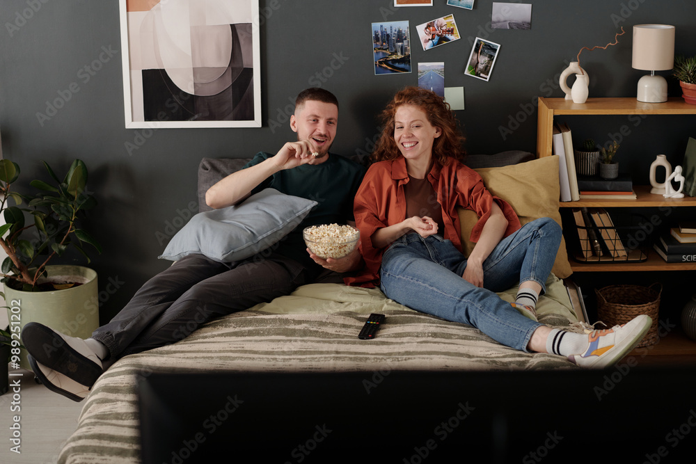 Young man explaining his girlfriend details of movie scene while both sitting on bed at home and having popcorn Young man explaining his girlfriend details of movie scene while both sitting on bed at home and having popcorn