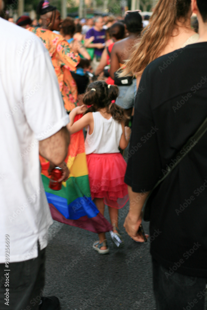 A young girl with pigtails and a red skirt is seen from behind, walking in a vibrant LGBTQ+ pride parade. She is holding part of a rainbow flag, symbolizing diversity, inclusion, and equality. The par A young girl with pigtails and a red skirt is seen from behind, walking in a vibrant LGBTQ+ pride parade. She is holding part of a rainbow flag, symbolizing diversity, inclusion, and equality. The par