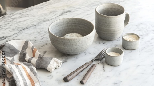 A grey ceramic bowl, two coffee mugs, and a small dip bowl sit on a marble countertop.  A fork and knife rest nearby, next to a kitchen towel. photo