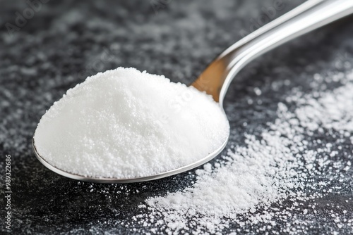 A silver teaspoon filled with granulated sugar, resting on a dark textured surface, with sugar granules scattered beside it.