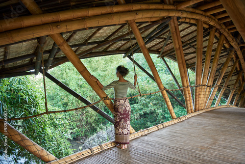Asia, Indonesia, Bali, Ubud, Mature Asian female tourist standing on bamboo bridge crossing small river, wearing asian style clothing