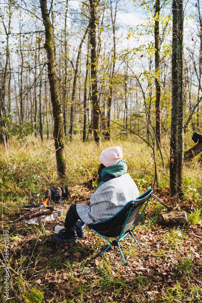 A woman is comfortably seated in a chair amidst the serene woods, close to a crackling campfire that provides warmth and light as she enjoys the beauty of nature around her A woman is comfortably seated in a chair amidst the serene woods, close to a crackling campfire that provides warmth and light as she enjoys the beauty of nature around her