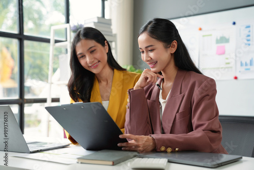 Two businesswomen are smiling while reviewing information on a clipboard during a meeting in the office