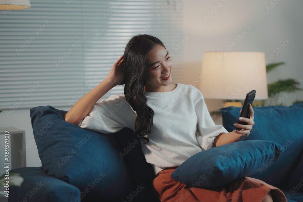Young asian woman happily scrolling on her phone in a cozy, modern living room at night, surrounded by stylish decor Young asian woman happily scrolling on her phone in a cozy, modern living room at night, surrounded by stylish decor