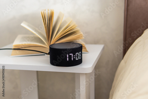 Round small smart speaker with digital clock and book on bedside table. Voice assistant at home photo