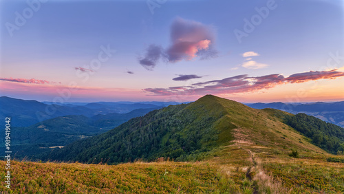 A vibrant sunset casts colorful hues over a mountain ridge, with rolling green hills stretching into the distance. Soft clouds float in a purple-pink sky, adding to the serene landscape