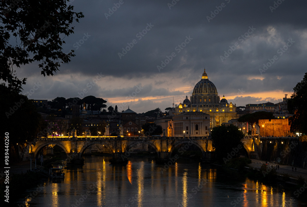 Fototapeta premium Evening view of St. Peter's Basilica across the Tiber River with illuminated buildings and dramatic clouds in Rome