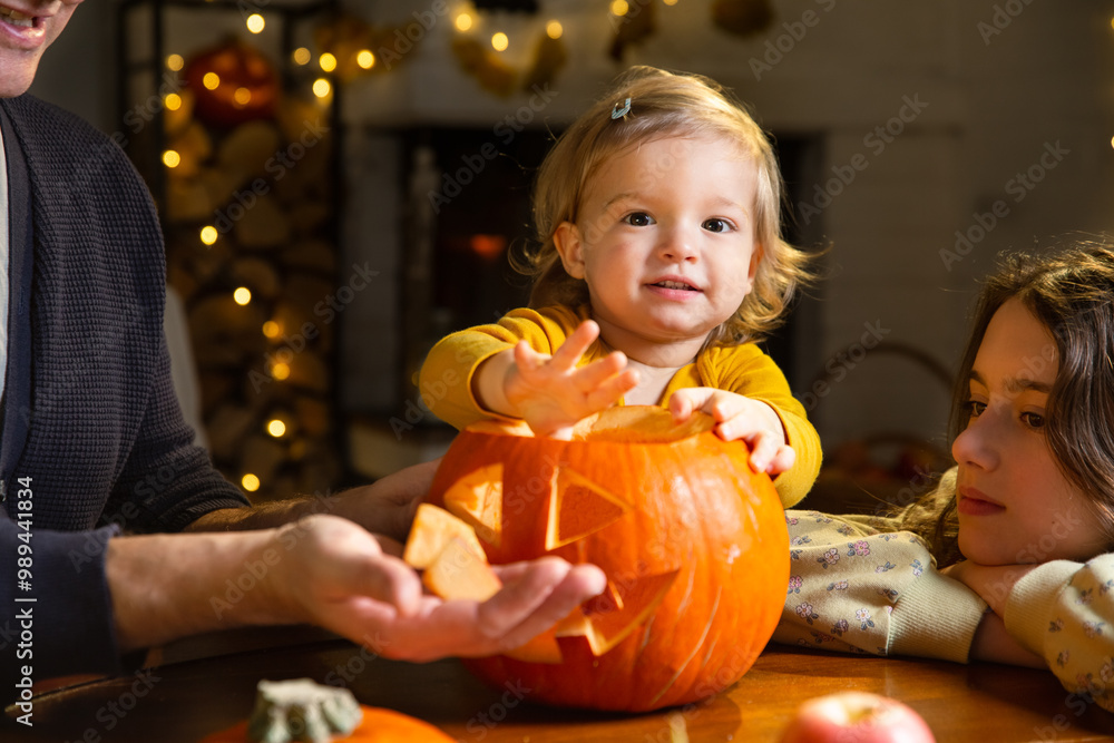 Fototapeta premium Father his toddler and daughter carving pumpkin jack o lantern, Halloween celebration at home. Fun craft and decoration at home. Happy children and their dad preparing for the holiday, decorating the