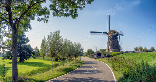 A winding road leads to a traditional windmill set against a clear blue sky. Lush green fields and trees line the sides of the road, creating a picturesque rural scene.