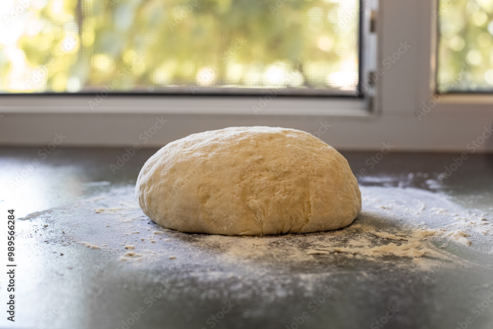 A lump of dough on a black table with scattered flour against the background of an open window. Kneaded dough for homemade baking. A lump of dough on a black table with scattered flour against the background of an open window. Kneaded dough for homemade baking.