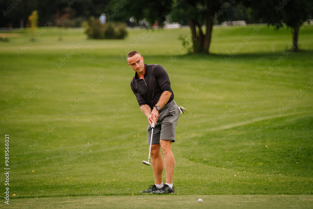 A focused golfer prepares to putt on a green golf course. He wears a black shirt and shorts, surrounded by trees and greenery. A focused golfer prepares to putt on a green golf course. He wears a black shirt and shorts, surrounded by trees and greenery.