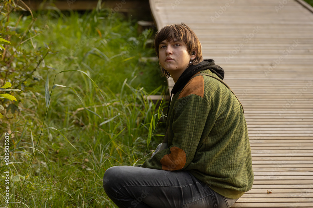 A teenage boy in a green hoodie sits by a wooden path surrounded by greenery. He gazes thoughtfully, with a quiet expression. The scene captures a moment of reflection in nature. A teenage boy in a green hoodie sits by a wooden path surrounded by greenery. He gazes thoughtfully, with a quiet expression. The scene captures a moment of reflection in nature.