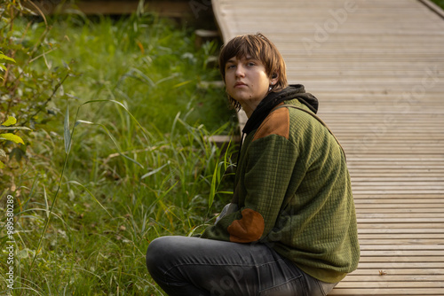 A teenage boy in a green hoodie sits by a wooden path surrounded by greenery. He gazes thoughtfully, with a quiet expression. The scene captures a moment of reflection in nature.