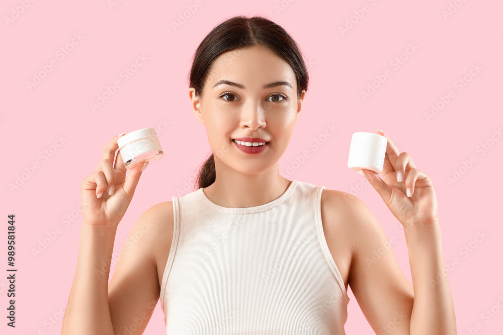 Pretty young woman with jars of facial cream on pink background