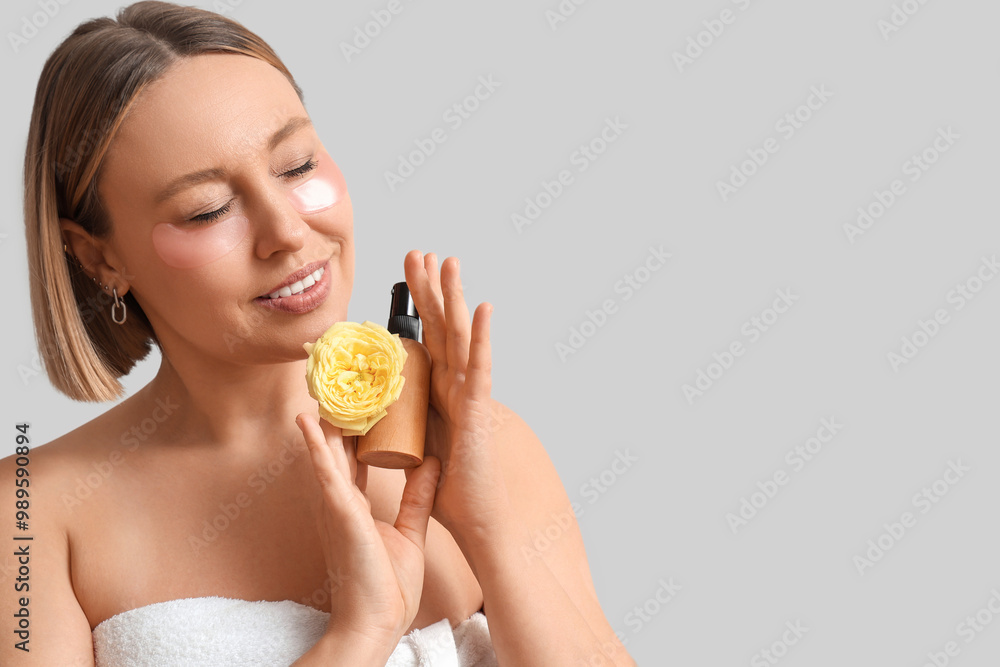 Beautiful mature woman with under-eye patches holding cosmetic product and flower on grey background, closeup Beautiful mature woman with under-eye patches holding cosmetic product and flower on grey background, closeup