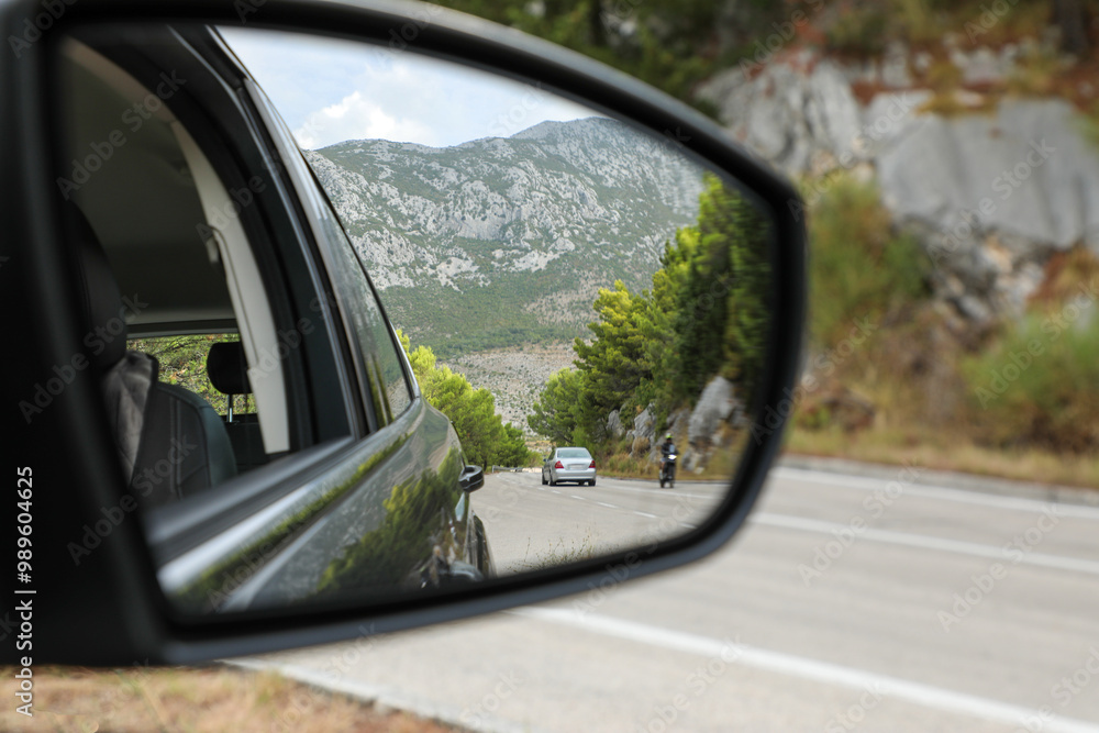 The driver looks into the side rearview mirror of the car and sees the road, high mountains and other road users. The driver looks into the side rearview mirror of the car and sees the road, high mountains and other road users.