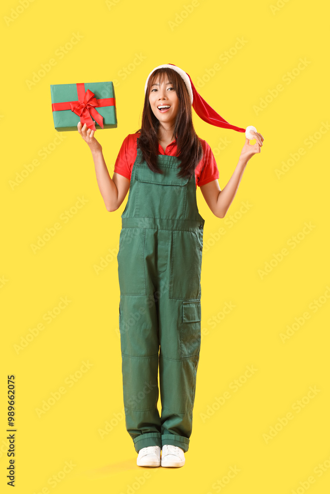 Female Asian worker in Santa hat with Christmas gift on yellow background Female Asian worker in Santa hat with Christmas gift on yellow background