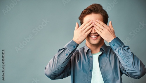 Smiling young man covering his eyes with both hands isolated on a blue background with large copy space to include text.