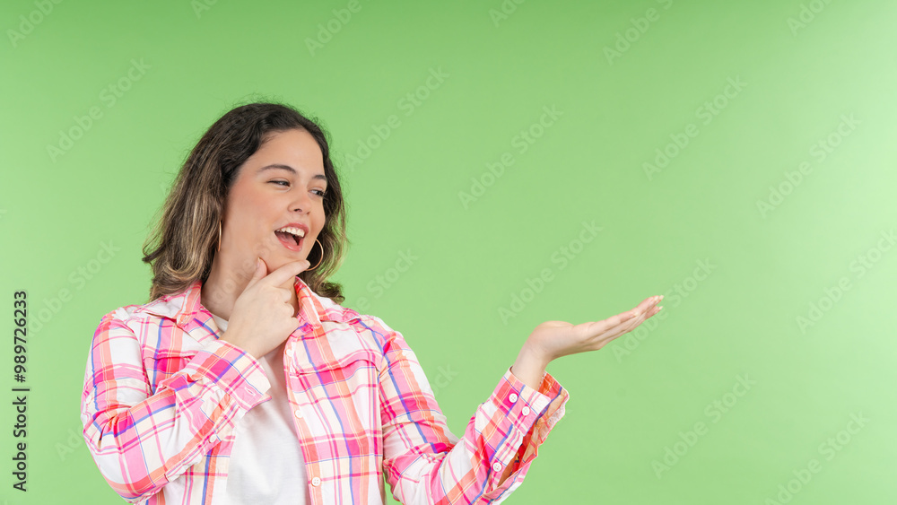 Young woman showing something with hand and touching chin on green background Young woman showing something with hand and touching chin on green background