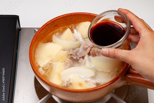 肉じゃがの調理写真。土鍋に油をひき、くし切りにしたタマネギとじゃがいも、豚肉を入れて炒めてだし汁出汁と醤油を加えて煮る