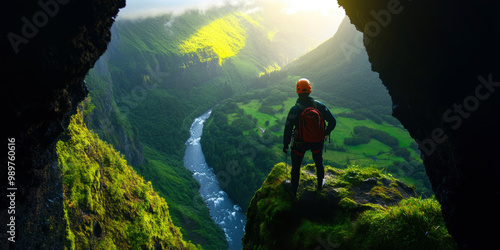 Standing atop a moss-covered cliff, a climber gazes out over a breathtaking valley, where a serene river meanders through lush greenery, bathed in golden morning light.