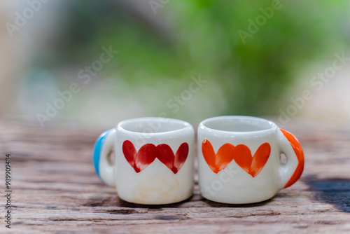 Two small, white ceramic mugs with colored heart designs sit side-by-side on a weathered wooden table. The background is blurred, with a soft green color suggesting a natural outdoor setting.
