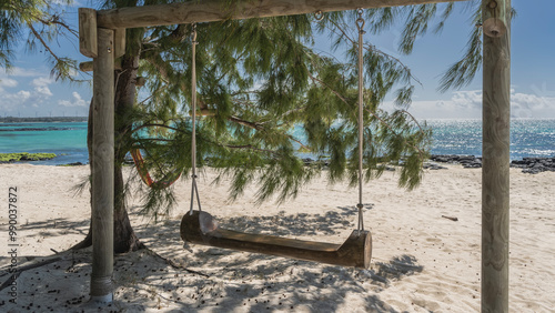 Wooden swings are suspended from ropes on log supports. The branches of the tree Casuarina cunninghamiana bent over the sandy beach. Shadows on the sand, fallen seeds. There is a turquoise ocean ahead photo