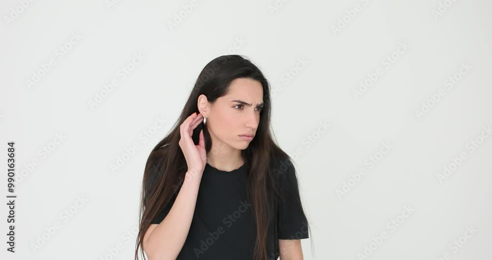 Curious woman with long loose dark hair holds hand next to ear trying to hear gossiping on white background. Female looks checking earring Curious woman with long loose dark hair holds hand next to ear trying to hear gossiping on white background. Female looks checking earring