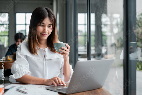 A young woman in a white blouse works on her laptop in a modern cafe, holding a cup of coffee and smiling.