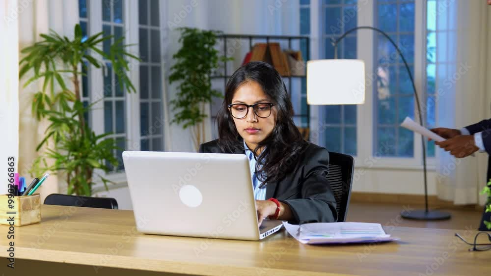 Young Indian manager focused on work at her desk, collaborating with a colleague who provides documents and instructions, offering guidance and support in a modern office environment Young Indian manager focused on work at her desk, collaborating with a colleague who provides documents and instructions, offering guidance and support in a modern office environment