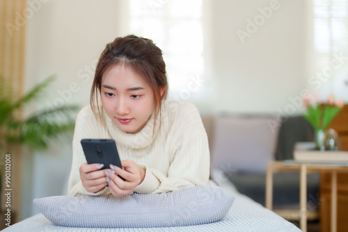 Young asian women in sweater lying on comfortable couch to enjoying with chatting social media on smartphone while relaxation and doing activity to spending time with slow life lifestyle at home