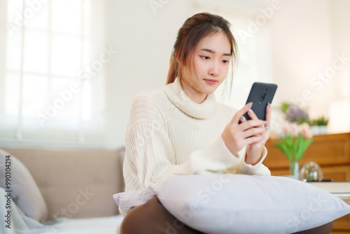 Young asian women in sweater enjoying with surfing entertainment on smartphone while sitting on comfortable couch to relaxation and doing activity for spending time with slow life lifestyle at home