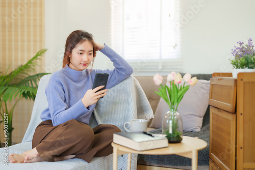 Young asian women in sweater using smartphone to watching on social media and entertainment while sitting on couch to relaxation and doing activity for spending time with slow life lifestyle at home